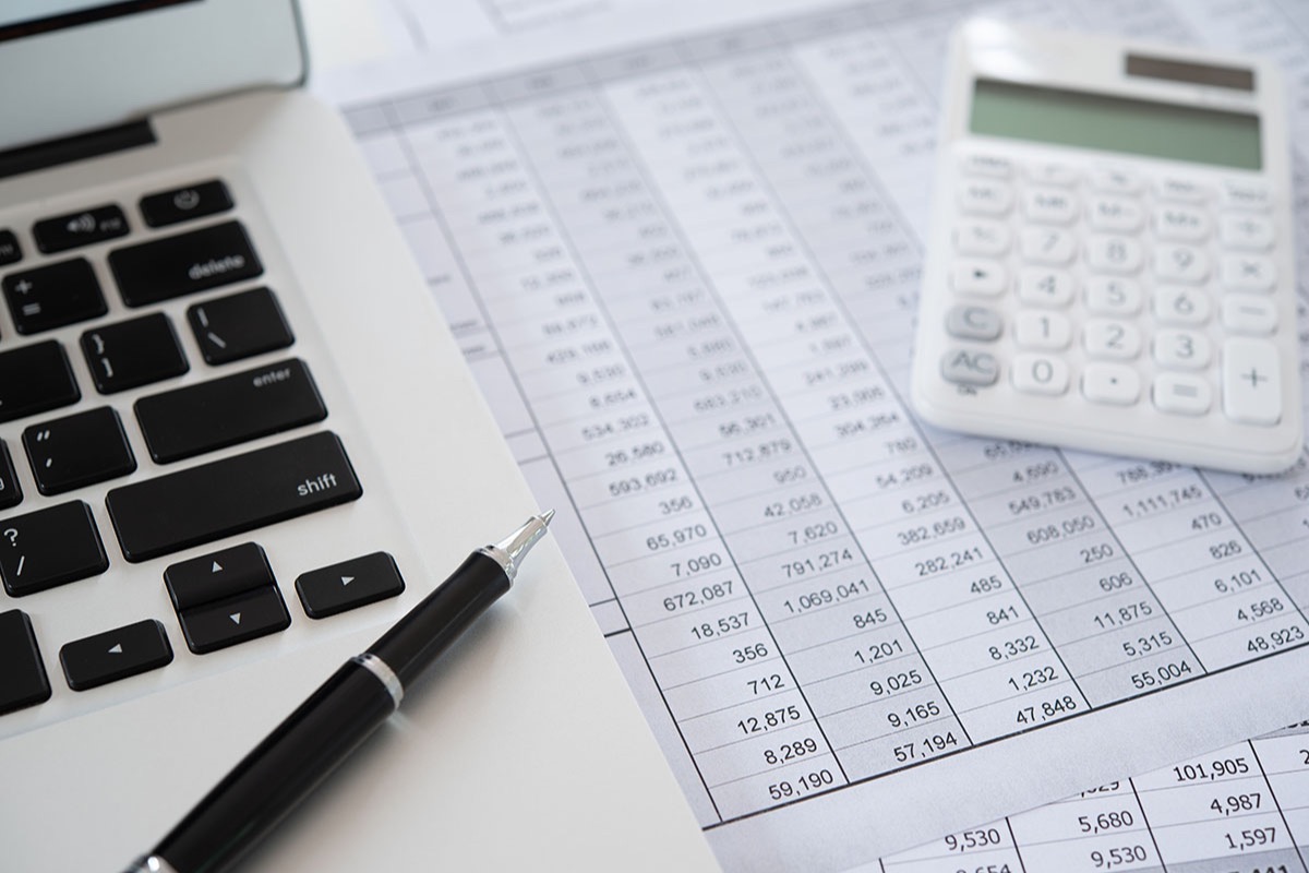 Laptop keyboard, black pen and white calculator resting on printed spreadsheets of financial figures on a desk