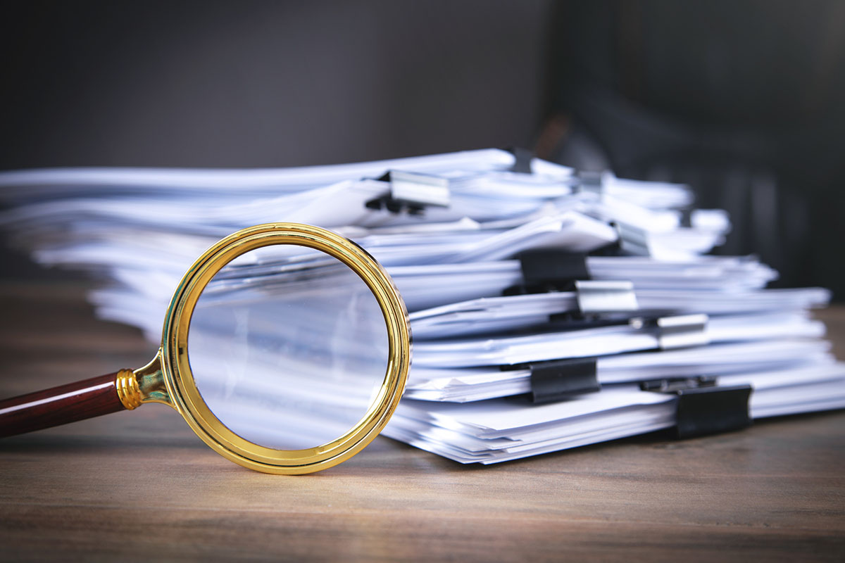 A brass magnifying glass leaning against a tall stack of paper documents held together with black binder clips on a wooden desk