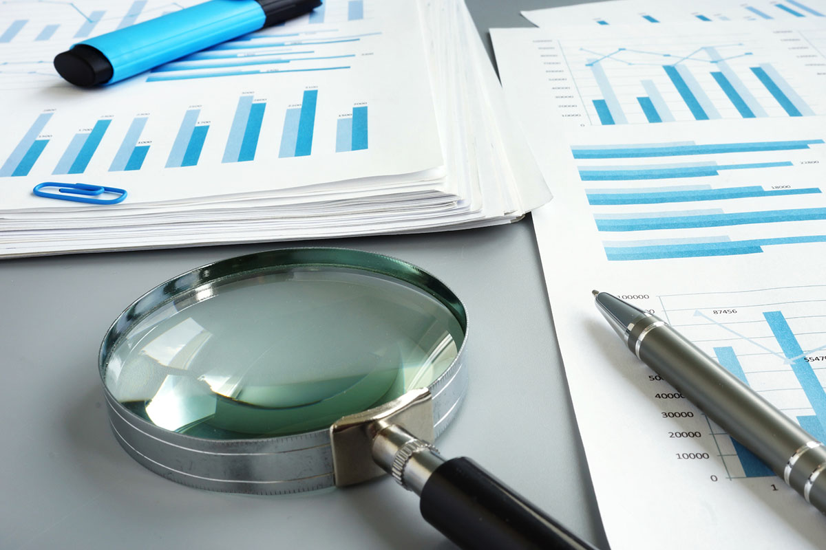 A large magnifying glass and a silver pen resting on printed financial reports covered in blue bar charts, beside a stack of papers and a blue highlighter on a grey desk