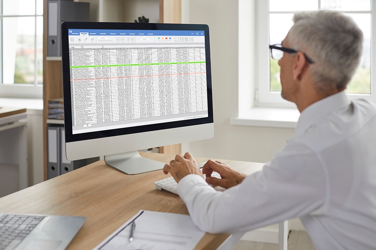 A gray-haired finance professional in a white shirt reviewing a large spreadsheet on a desktop monitor in a bright office, with a laptop and printed documents on the desk