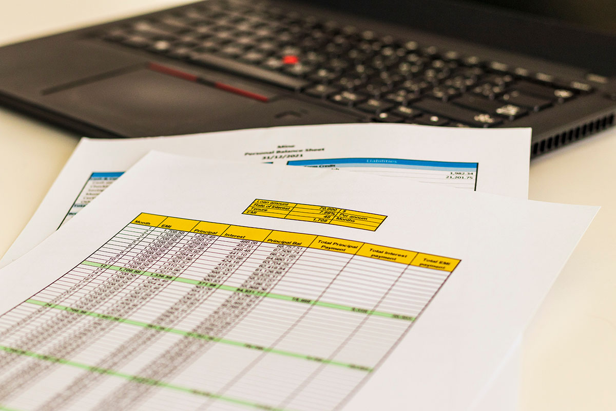 Printed spreadsheet pages with yellow header rows and columns of payment figures on a white desk in front of a black laptop keyboard